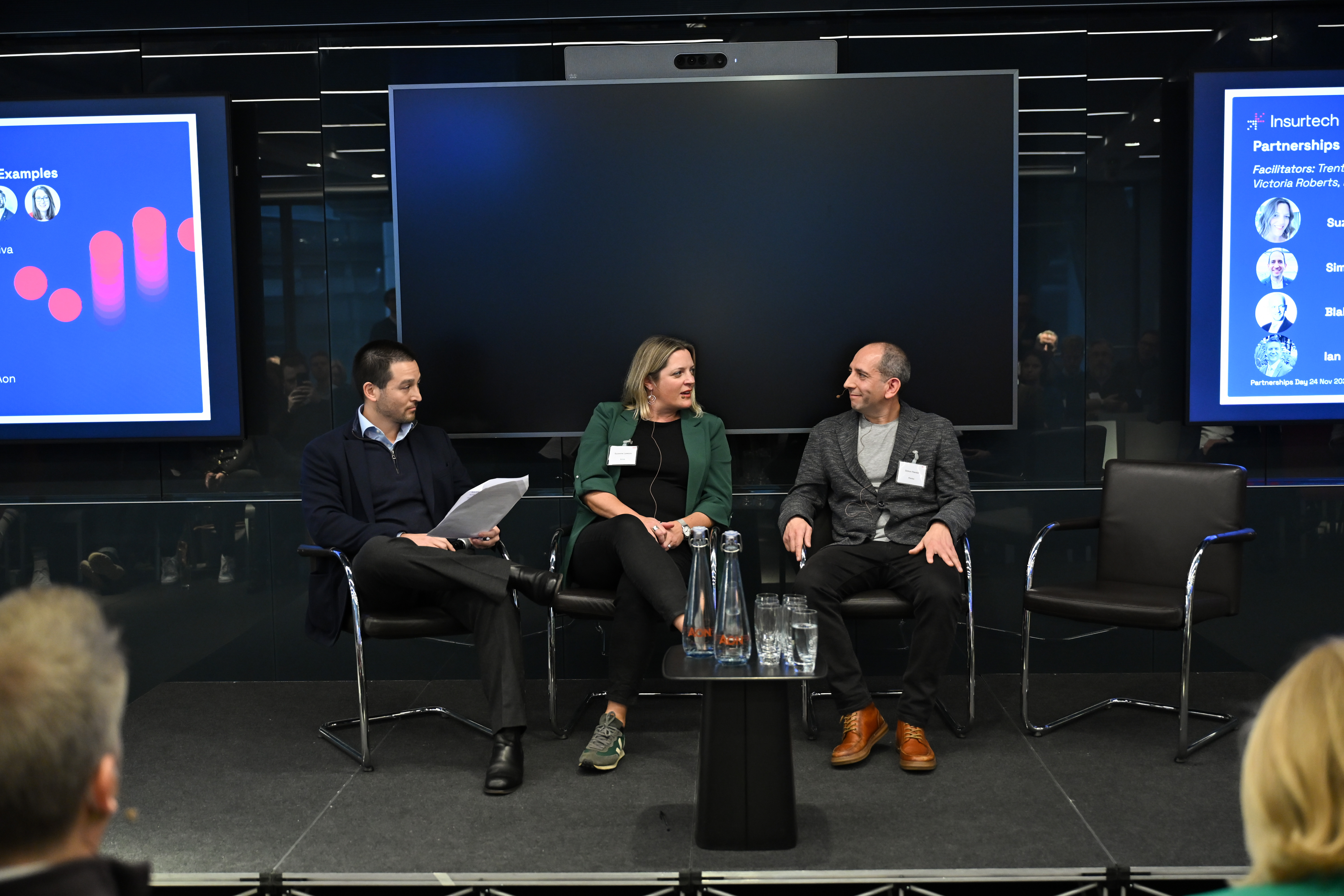 Woman in business attire, flanked by two men, all sitting on stage